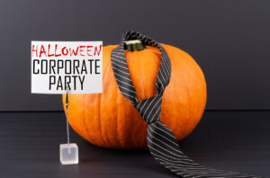 An orange pumpkin with a tie wrapped around it sitting next to a sign with "Halloween Corporate Party" printed on it
